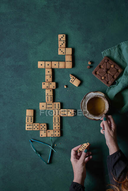 Top view of crop anonymous person eating sweet domino shaped cookies and drinking hot coffee on green background with eyeglasses — Stock Photo
