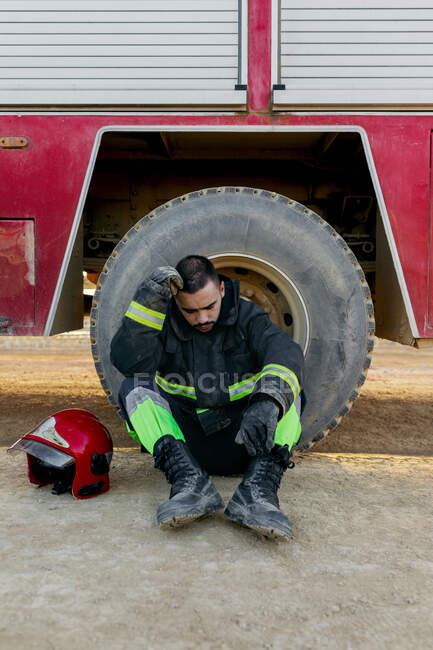 Exhausted male firefighter wearing uniform sitting on ground near fire engine near red helmet while leaning his head on hand — Stock Photo
