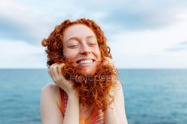 Joven pelirroja alegre haciendo pose infantil con el pelo rizado mientras disfruta de la libertad en la orilla del mar con los ojos cerrados - foto de stock
