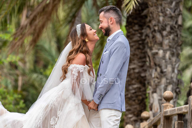 Couple marié dans des tenues de mariage debout sur la passerelle en bois avec rampe tout embrassé et en regardant les uns les autres près des palmiers verts et des plantes dans le jardin en journée d'été — Photo de stock