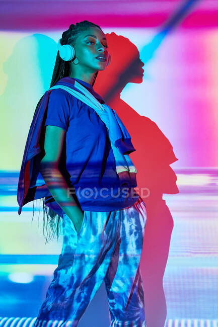 Бічний погляд на модну молоду домініканську жінку тисячолітня з довгими африканськими плечима, що стоять на підлозі і дивляться на неї, слухаючи музику в навушниках в кімнаті з барвистим геометричним освітленням. — стокове фото