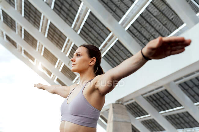 Mujer decidida en ropa deportiva con brazos extendidos haciendo ejercicio de yoga en la calle cerca del panel fotovoltaico moderno durante el entrenamiento en la ciudad - foto de stock
