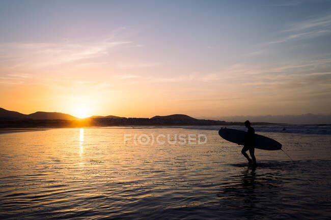 Vista laterale della silhouette anonima dell'atleta con la tavola da surf che cammina nell'oceano increspato contro Famara Mountain al tramonto a Lanzarote Spagna — Foto stock