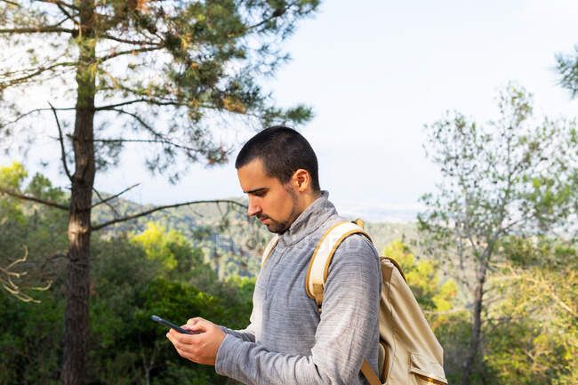 Vista lateral del joven viajero masculino étnico barbudo concentrado en ropa casual y mensajería de mochila en un teléfono inteligente de pie en un exuberante bosque verde durante el trekking en el valle montañoso - foto de stock