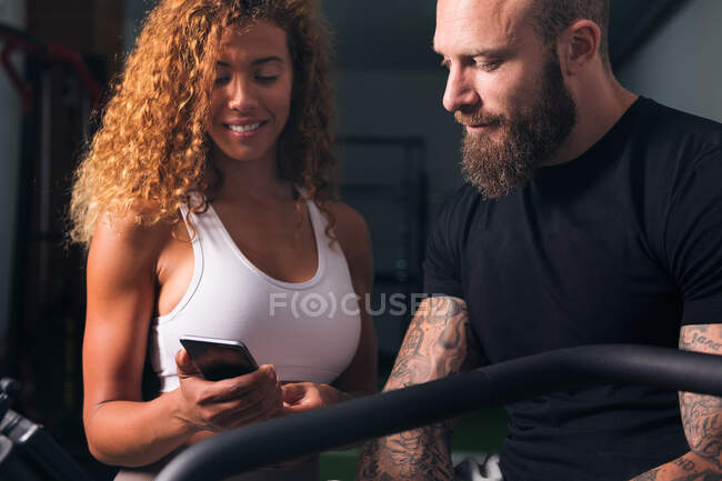 Mujer con cabello rizado en top blanco demostrando smartphone a hombre con tatuajes en la habitación - foto de stock