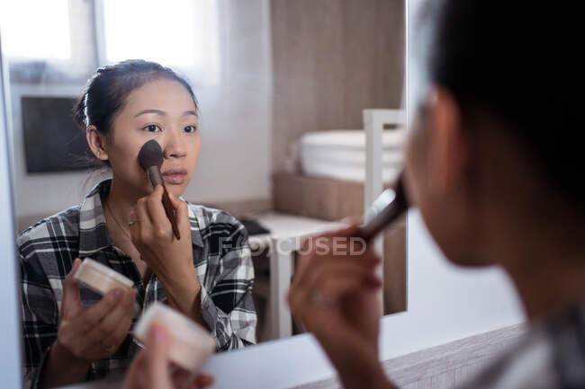 Бічний вид на молоду азіатську жінку в повсякденному одязі, що використовує порошок, стоячи перед дзеркалом у світлій спальні. — стокове фото