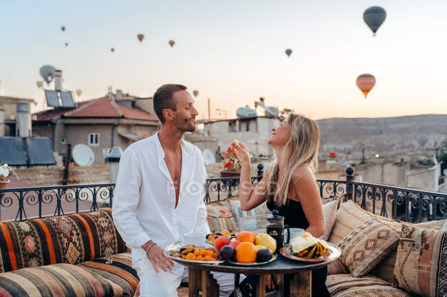 Couple holding hands and looking at each other while enjoying romantic dinner and feeding each other with fruits on terrace with hot air balloons on background in Cappadocia — Stock Photo