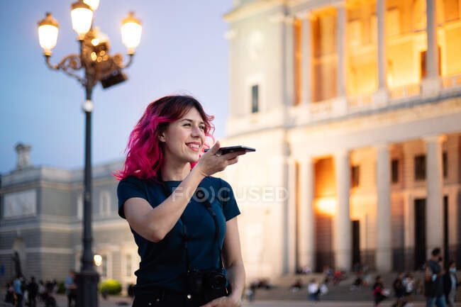 Веселая женщина с розовыми волосами записывает голосовое сообщение, стоя на улице с фонариком рядом с классическим светящимся зданием в городе — стоковое фото