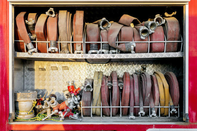 Набор различных типов скрученных пожарных шлангов и различных типов адаптеров, расположенных на двух полках в пожарной машине при ярком солнечном свете днем — стоковое фото