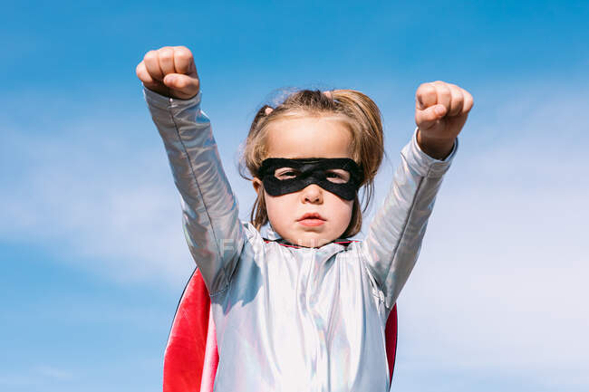De baixo da menina pequena em traje de super-herói levantando punhos estendidos para mostrar poder enquanto estava contra o céu azul claro — Fotografia de Stock