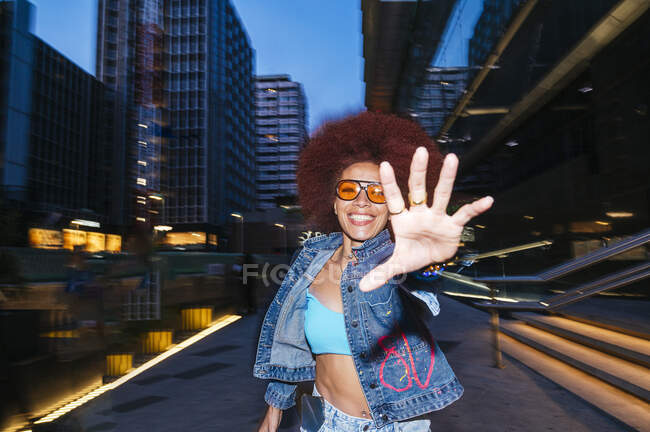 Fröhliche Frau im trendigen Outfit mit Sonnenbrille läuft und reicht der Kamera die Hand auf der dunklen Straße mit modernen Gebäuden — Stockfoto
