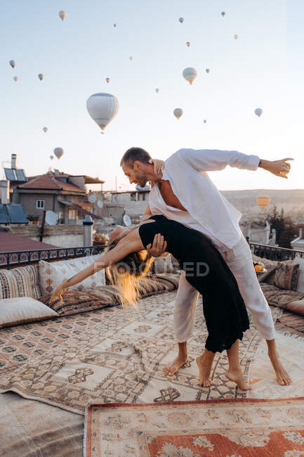 Cuerpo completo de pareja descalza bailando juntos en la terraza de la azotea contra globos de aire caliente volando en cielo despejado - foto de stock