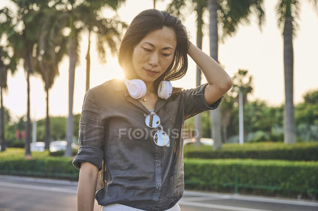 Mujer asiática seria con auriculares blancos modernos mirando hacia abajo mientras está de pie cerca de la carretera en la calle de la ciudad con árboles verdes - foto de stock