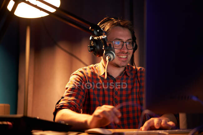 Vista lateral de un joven enfocado en camisa a cuadros y anteojos usando computadora y hablando en micrófono mientras graba podcast en estudio oscuro - foto de stock