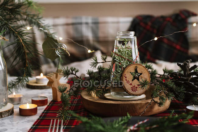 Ajuste de mesa de Navidad con corona en el plato, adornos decorativos de madera y mantel a cuadros rojo con luces amarillas en el fondo - foto de stock