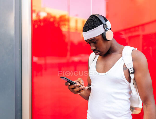 Focused African American male in white clothes listening to music in wireless headphones and messaging on mobile phone while standing against red wall in city — Stock Photo