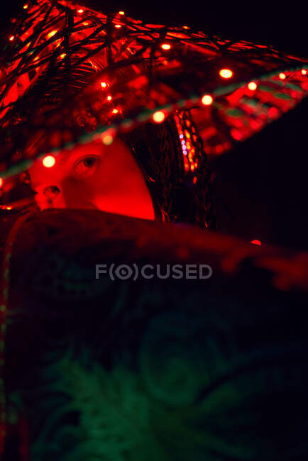 Enigmatic female in creative traditional outfit and Vietnamese headwear with red illumination standing in dark studio on black background during performance — Stock Photo