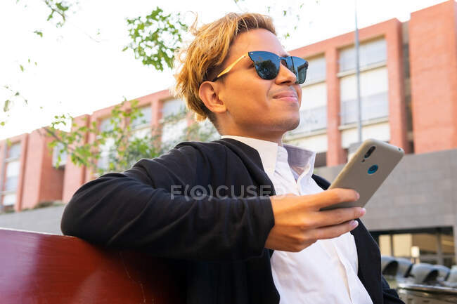 Joven hombre alegre en gafas de sol y elegante traje de mensajería de texto en el teléfono celular mientras está sentado en la calle cerca del edificio en la ciudad - foto de stock