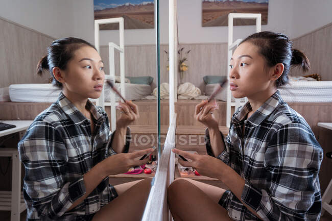 Vista laterale della giovane donna asiatica in abiti casual applicando polvere mentre in piedi davanti allo specchio in camera da letto leggera — Foto stock