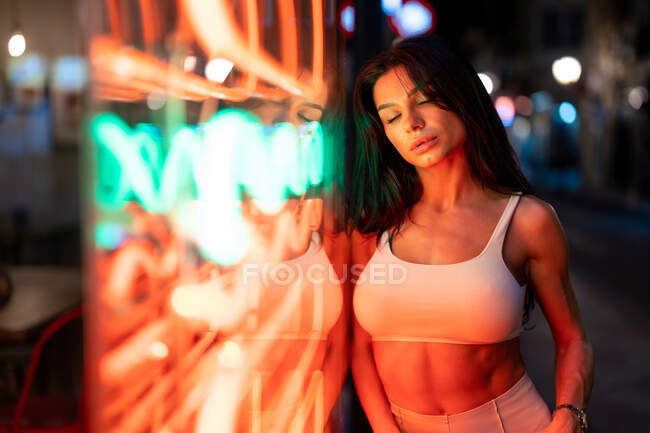 Charming female in white top closed eyes while standing near building with glowing lights in evening time on street — Stock Photo
