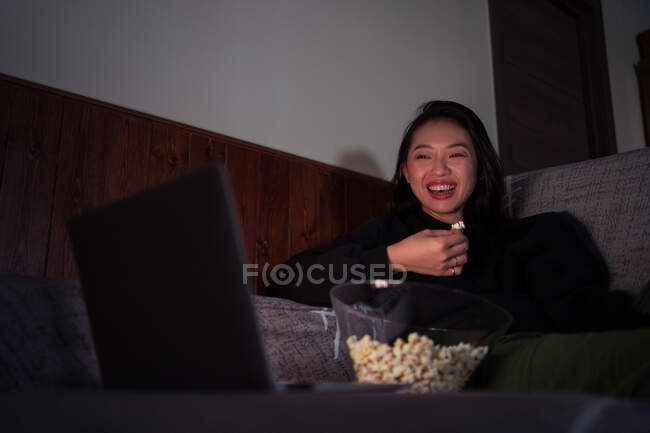 Jovem mulher asiática alegre vestindo roupas casuais sentado no sofá e comendo pipocas em casa assistindo filme no laptop — Fotografia de Stock