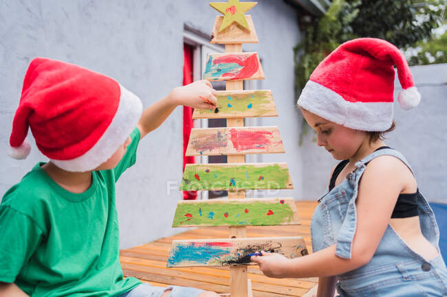 Side view of concentrated kids in casual wear painting wooden Christmas tree together in light room during festive holiday preparation — Stock Photo