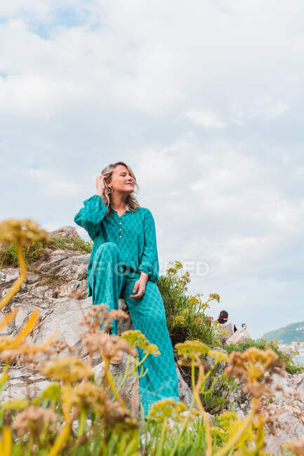 Full body of positive female in stylish outfit adjusting hair and sitting on rocks with green plants in San Sebastian in Spain against cloudy blue sky in daytime — Stock Photo
