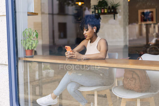 Através da janela de jovem fêmea étnica com cabelos negros afro na roupa da moda e verdadeiros fones de ouvido sem fio felizmente ao ter uma conversa em vídeo no smartphone no café moderno — Fotografia de Stock