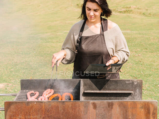 Chef femenino concentrado con pinzas en delantal cocinando salchichas en la parrilla mientras está de pie en el campo cubierto de hierba en el campo - foto de stock