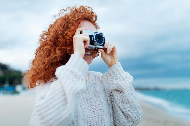 Donna dai capelli rossi positiva in maglione lavorato a maglia che scatta foto sulla macchina fotografica retrò sulla costa del mare — Foto stock