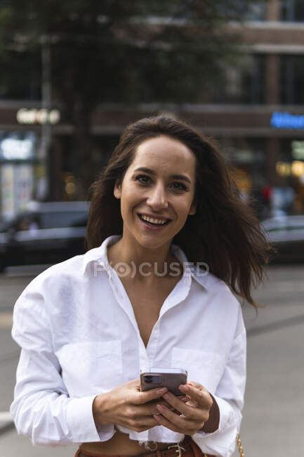 Mulher confiante em roupa elegante usando smartphone enquanto está de pé na rua da cidade perto da estrada com carros — Fotografia de Stock