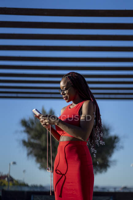 Side view of African American female with braids in trendy red outfit standing and browsing smartphone on street against residential building in city in sunlight — Stock Photo