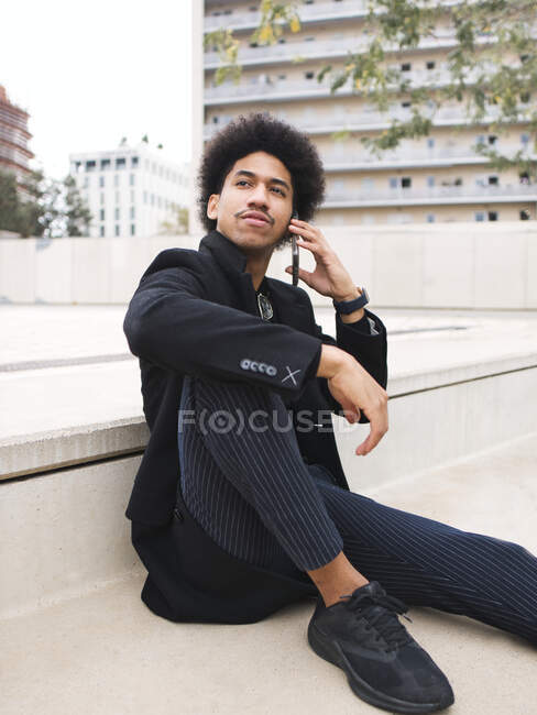 Young black man with dark Afro hair in trendy outfit talking smartphone on city square — Stock Photo