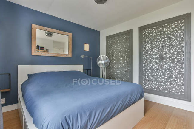 Cama confortável com cobertor azul colocado no quarto elegante com ventilador e elementos decorativos criativos na parede no apartamento moderno — Fotografia de Stock