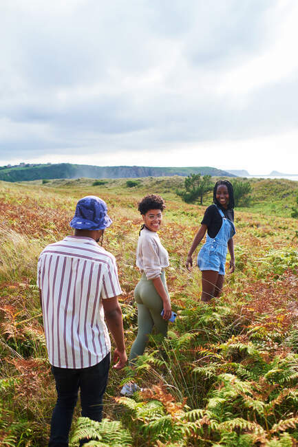Visão traseira de amigos afro-americanos positivos em roupas casuais andando entre a grama levando a penhasco rochoso e beira-mar no dia de verão — Fotografia de Stock