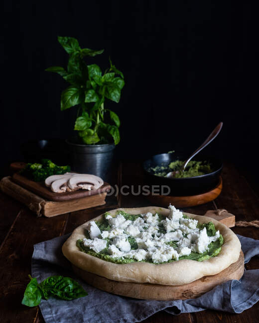 Appetizing vegetarian uncooked pizza with Mozzarella cheese and green pesto sauce placed on table with basil leaves in dark room — Stock Photo