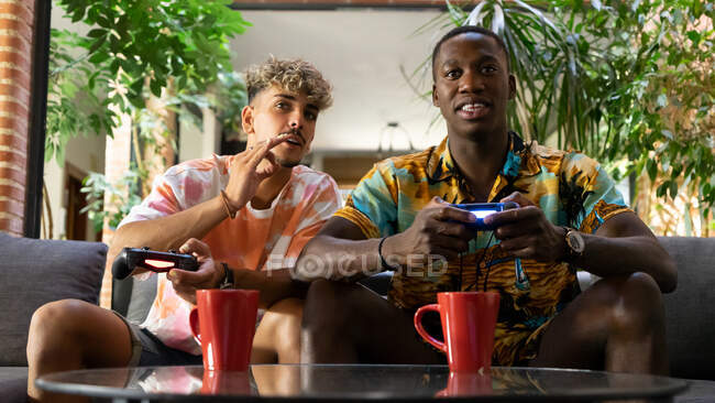 Diversos amigos do sexo masculino com gamepads beber bebidas enquanto sentado no sofá e jogar videogame juntos na sala de estar com plantas verdes — Fotografia de Stock