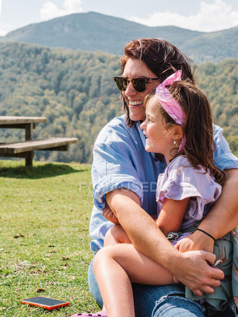 Madre positiva en gafas de sol con linda hija en vueltas sentada en terreno herboso contra la montaña con árboles verdes en la naturaleza - foto de stock