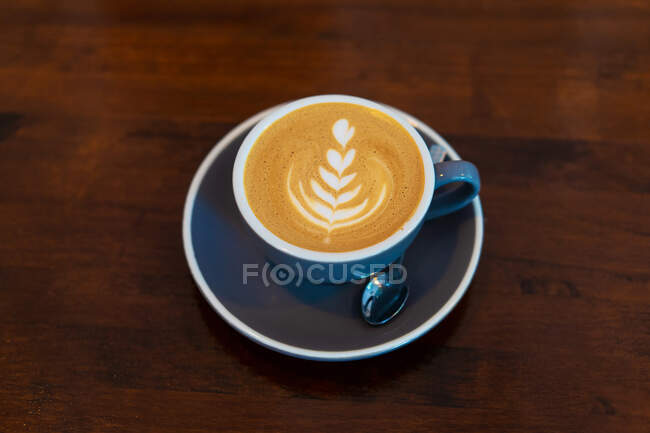 Сверху чашка горячего кофе с искусством латте на тарелке с чайной ложкой подается на деревянном столе в светлом кафе — стоковое фото