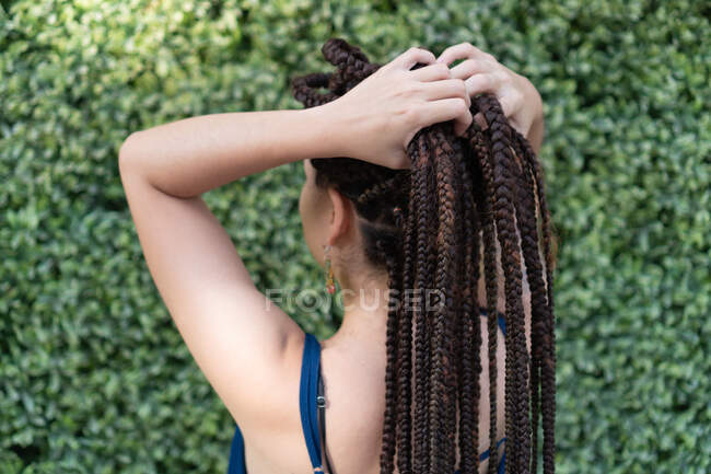 Вид сзади на анонимную латиноамериканку с татуировкой и длинными черными плетеными волосами во время стояния и размещения волос с руками рядом с зелеными растениями — стоковое фото
