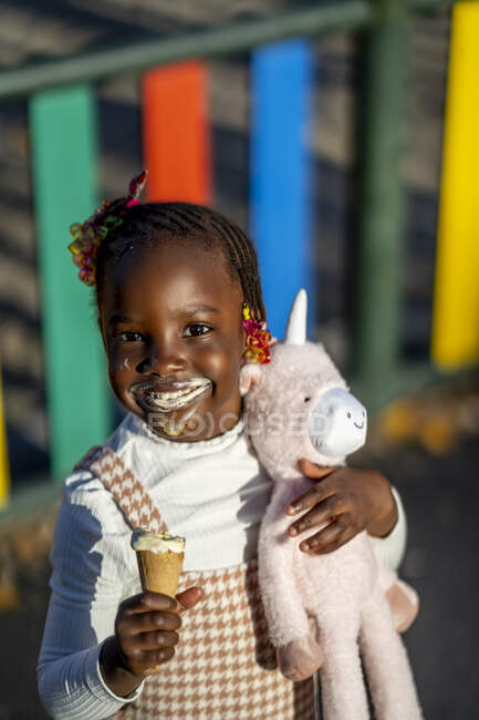 Cheerful African American girl with hairstyle standing eating ice cream and hugging teddy on street in sunny day — Stock Photo