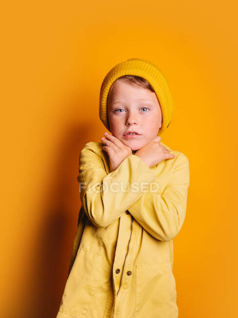 Niño poco emocional en impermeable de moda y gorro sombrero de pie con los brazos cruzados y mirando a la cámara contra el fondo amarillo en el estudio - foto de stock