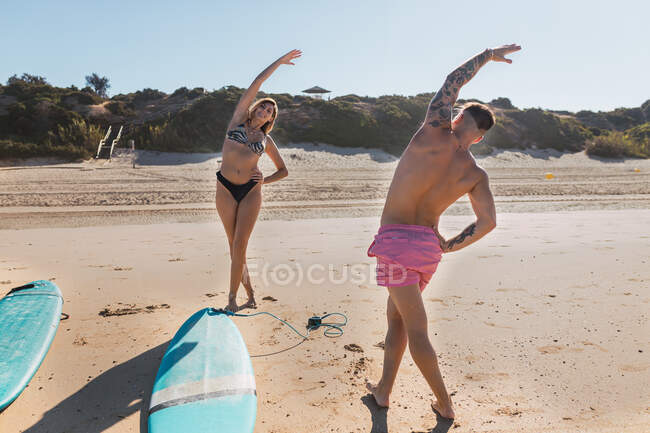 Full body of sportive couple in swimwear looking at each other while stretching body on sunny sandy beach with surfboards — Stock Photo