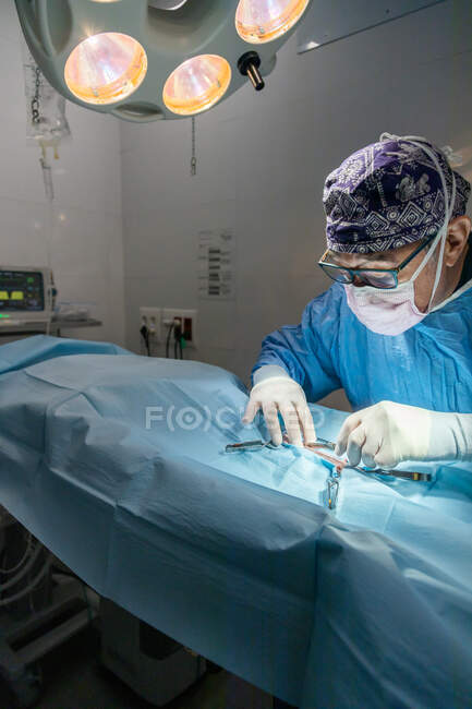 Profissional cirurgião masculino sênior em máscara e uniforme fazendo operação sob lâmpada na sala de cirurgia — Fotografia de Stock