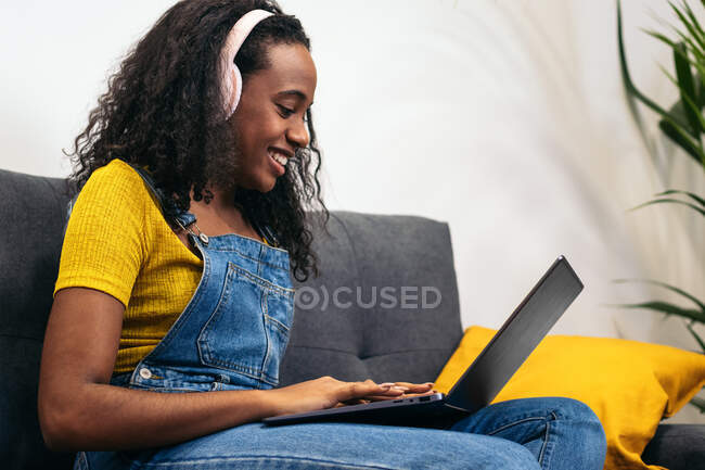 Mulher negra sorridente em macacão jeans com fones de ouvido sentados no sofá e usando laptop em casa — Fotografia de Stock