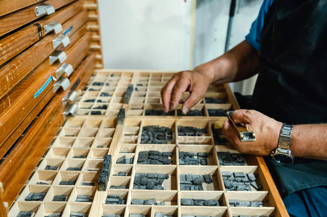 From above of crop concentrated senior male artisan in apron and eyeglasses choosing printing press letters from wooden box during work in traditional atelier — Stock Photo