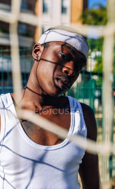Grave atleta afroamericano maschio in activewear bianco in piedi vicino alla rete sul parco giochi in estate e guardando la fotocamera — Foto stock
