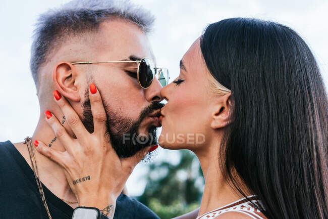 Side view of female with dark hair and red nails touching face of boyfriend in sunglasses while kissing with closed eyes against green trees on street in daylight — Stock Photo