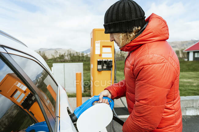 Side view of thoughtful male in warm outwear and hat standing with charging power to electric vehicle in hand at gas station in sunny day — Stock Photo