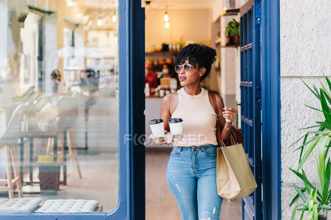 Positive young African American female with curly hair in casual clothes while leaving modern cafe with takeaway coffee cups and paper bags — Stock Photo